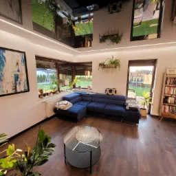 Jasne wnętrze salonu z ciemną podłogą, niebieskim narożnikiem, szklanym stolikiem kawowym i roślinami doniczkowymi, widok na ogród przez okna.