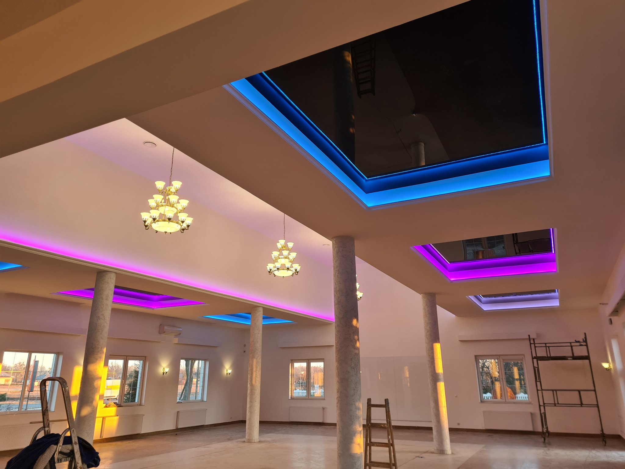Wnętrze przestronnego pomieszczenia z kolumnami, oświetleniem LED w kolorach niebieskim i fioletowym w suficie oraz żyrandolami; widoczne drabiny i rusztowanie wskazują na trwające prace...
