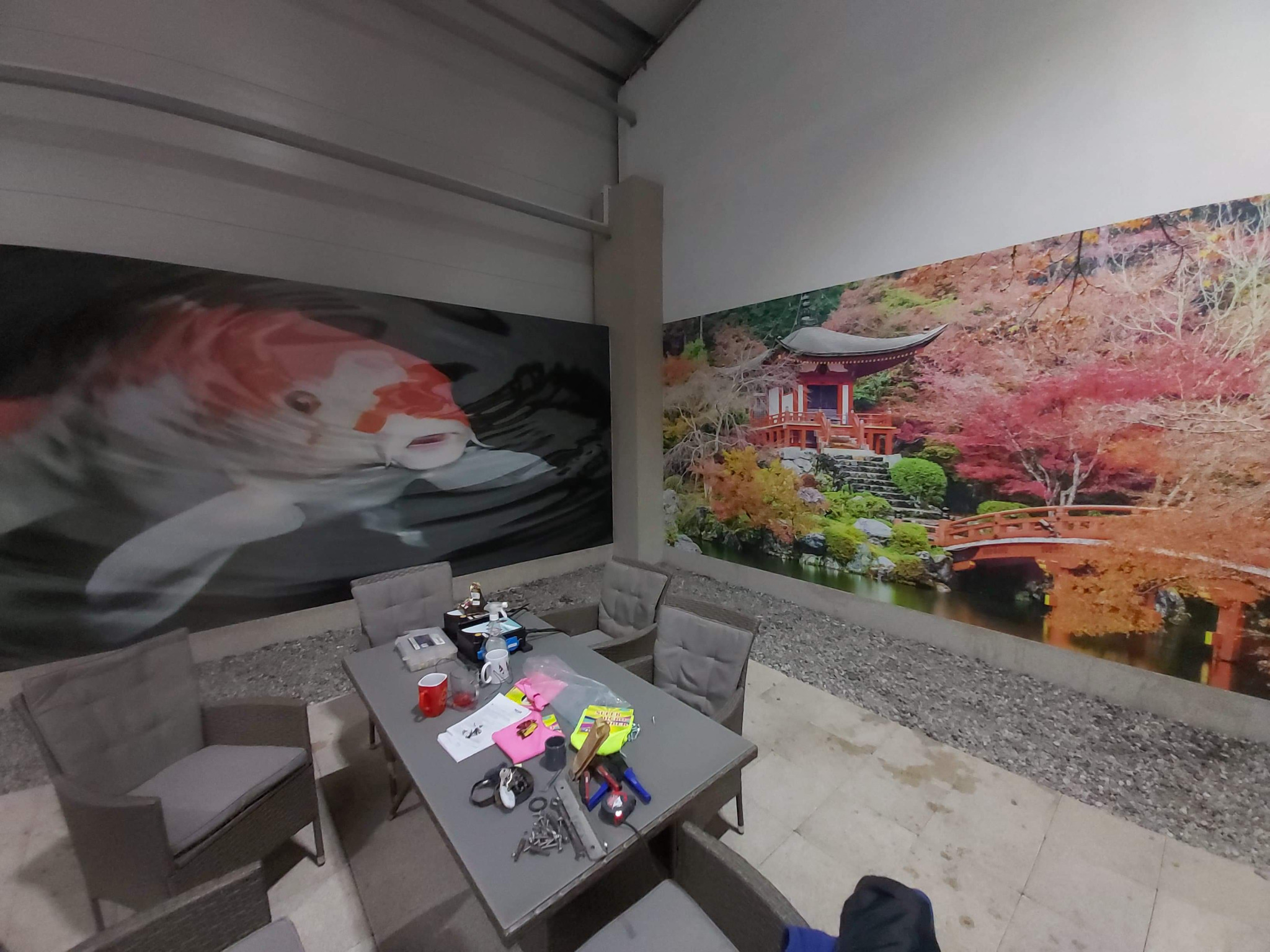 Wnętrze pomieszczenia z fototapetami przedstawiającymi japoński ogród i karpia koi, stół z narzędziami i krzesłami.