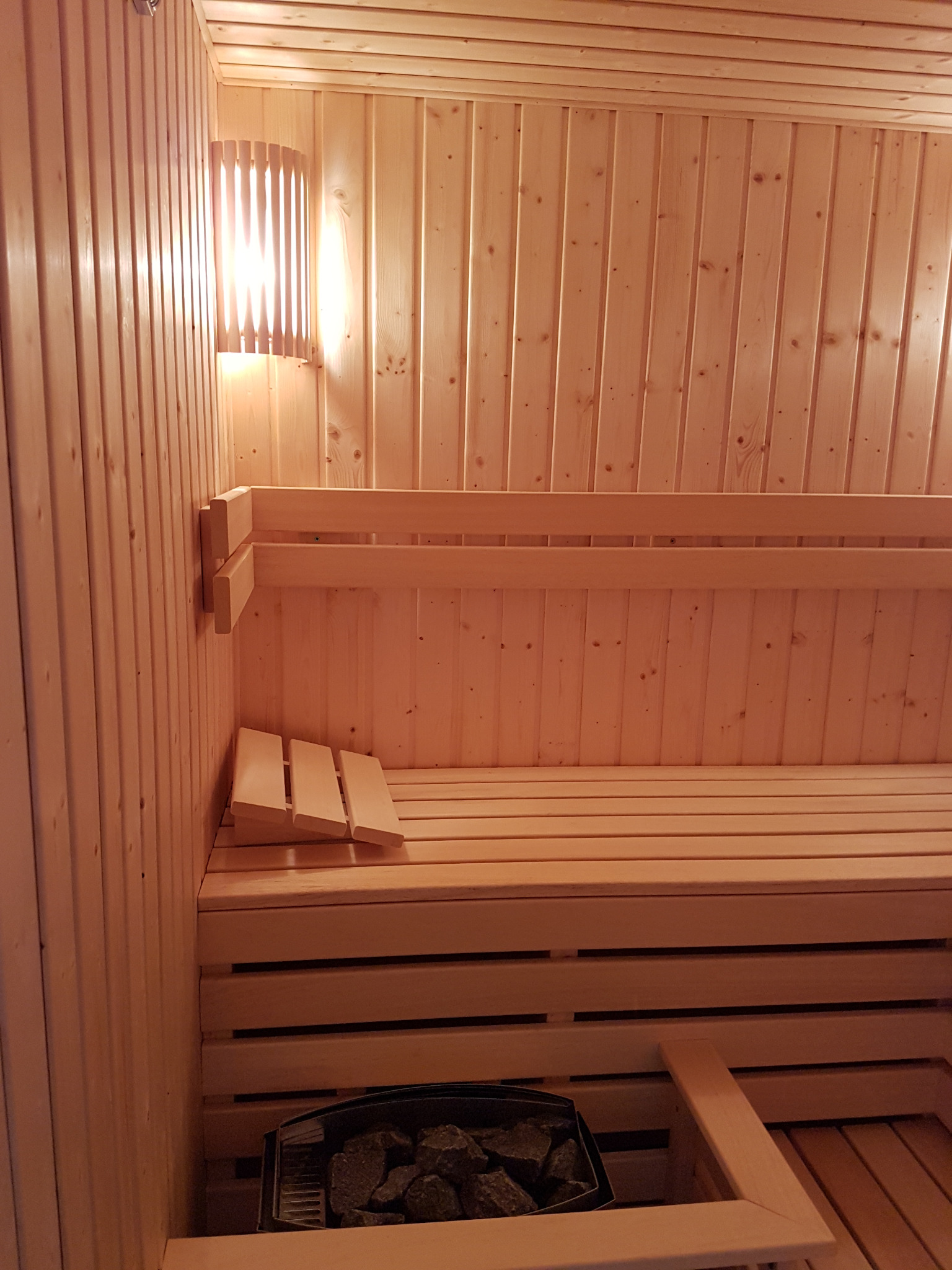 Wnętrze sauny wyłożone jasnym drewnem, z drewnianymi ławkami, oświetlone lampą ścienną z drewnianymi listwami, widoczne kamienie w piecu.