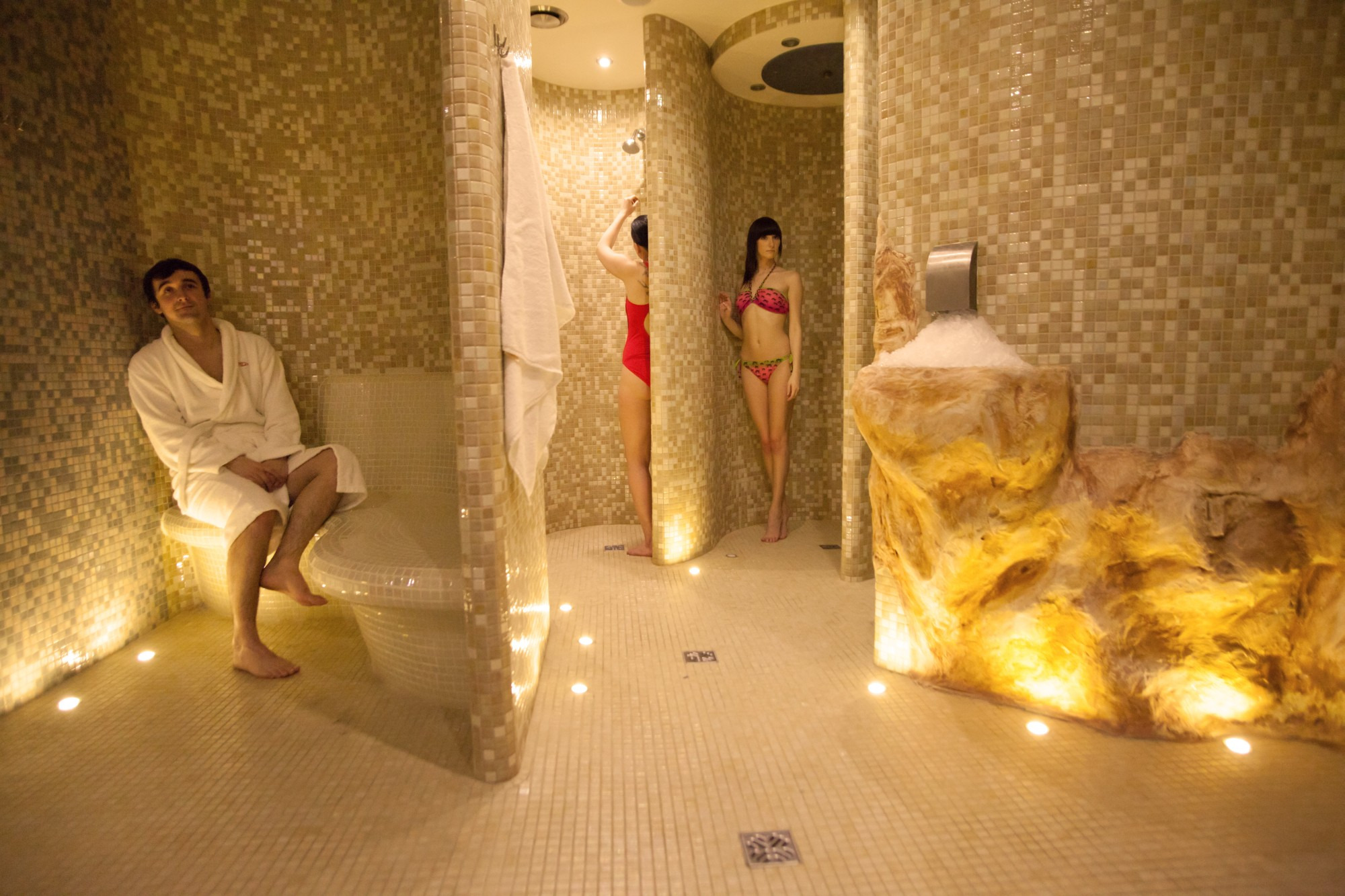 Wnętrze luksusowego spa z mozaikowymi ścianami, oświetleniem punktowym w podłodze, mężczyzną w szlafroku siedzącym na mozaikowej ławie oraz dwiema kobietami w strojach kąpielowych w kabinach...