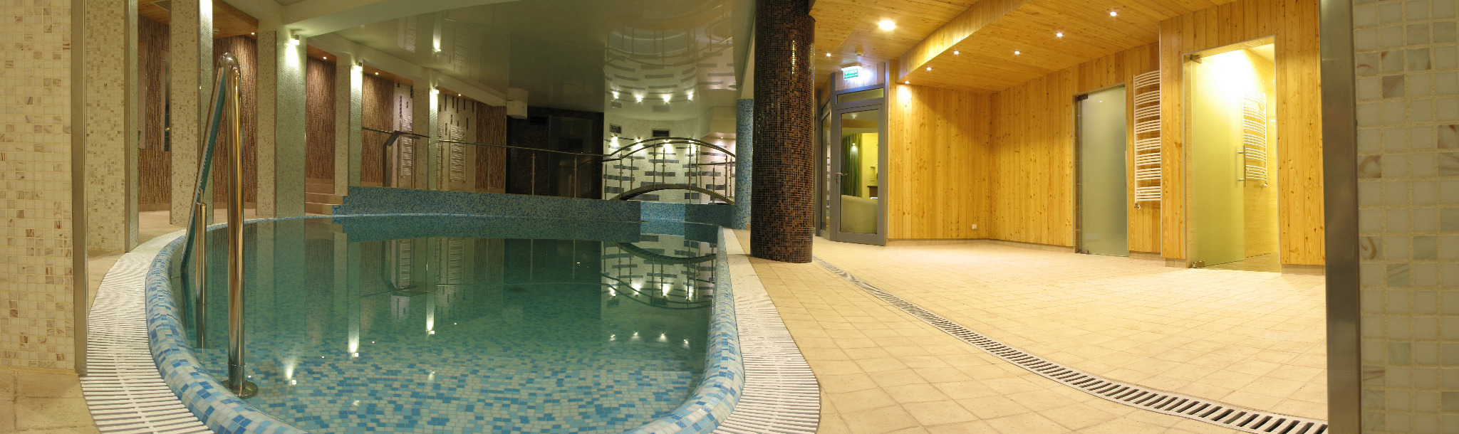 Kryty basen z mozaikową obudową i metalową barierką, widok na drewnianą saunę z przeszklonymi drzwiami i grzejnikiem drabinkowym.