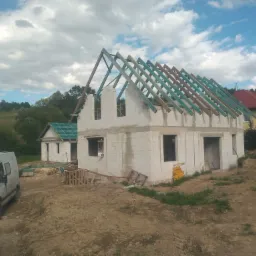 Budowa domu z białych bloczków w Sanoku, widoczne ściany i konstrukcja dachu z zielonymi elementami, na pierwszym planie biały bus.