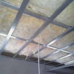 Montaż izolacji z wełny mineralnej w metalowym stelażu podwieszanego sufitu, widoczne kable instalacji elektrycznej.