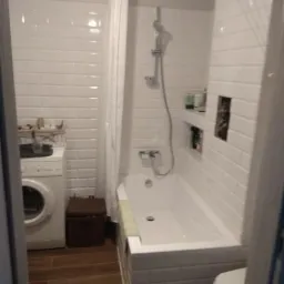 Biała łazienka z wanną obudowaną płytkami imitującymi cegłę, pralką i brązową skrzynią na tle ściany wyłożonej białymi płytkami typu metro.