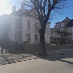 Dwa budynki mieszkalne z elewacjami w odcieniach szarości i zieleni, oddzielone drzewem bez liści, widoczne ogrodzenie i fragment ulicy w słoneczny dzień.