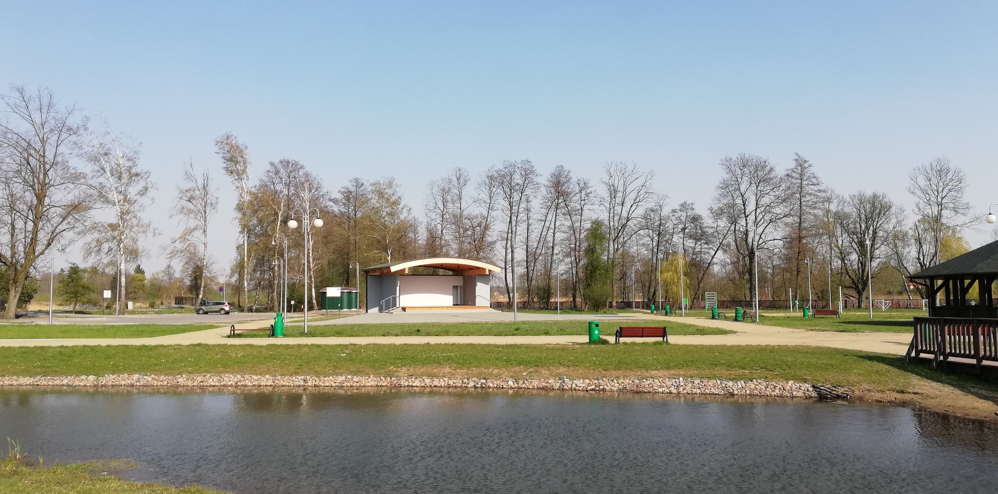 Amfiteatr nad stawem w parku miejskim. Scena z zadaszeniem, ławki, oświetlenie parkowe. W tle drzewa bez liści i błękitne niebo. Woda odbija otoczenie.