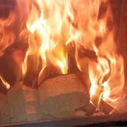 Płonące brykiety drzewne wewnątrz kominka z czarną rączką do otwierania drzwiczek, widoczne płomienie i żar