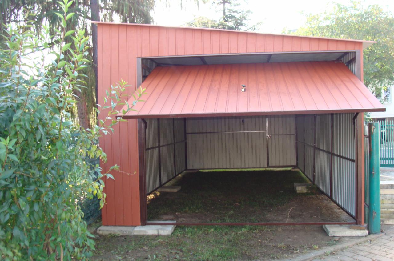 Otwarty garaż blaszany z podnoszoną bramą w kolorze ceglastym, widok na wnętrze z ziemią i trawą, otoczenie drzew i zieleni.