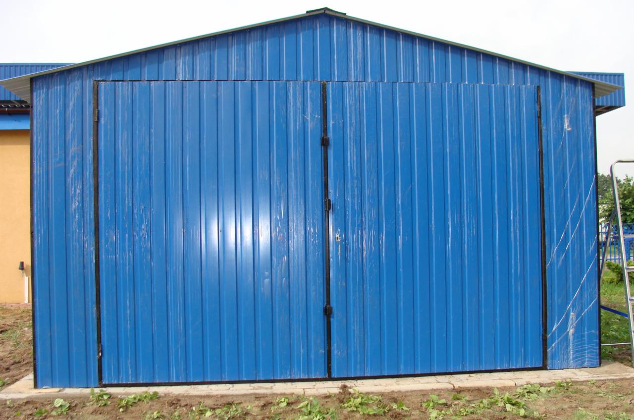 Niebieski garaż blaszany z dwuskrzydłowymi drzwiami, widoczne ślady folii ochronnej na poszyciu, teren wokół jeszcze niezagospodarowany.