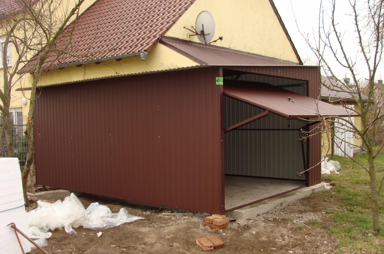 Brązowy garaż blaszany z uchyloną bramą segmentową, widoczne elementy konstrukcyjne i betonowa podstawa, teren wokół w trakcie prac wykończeniowych.
