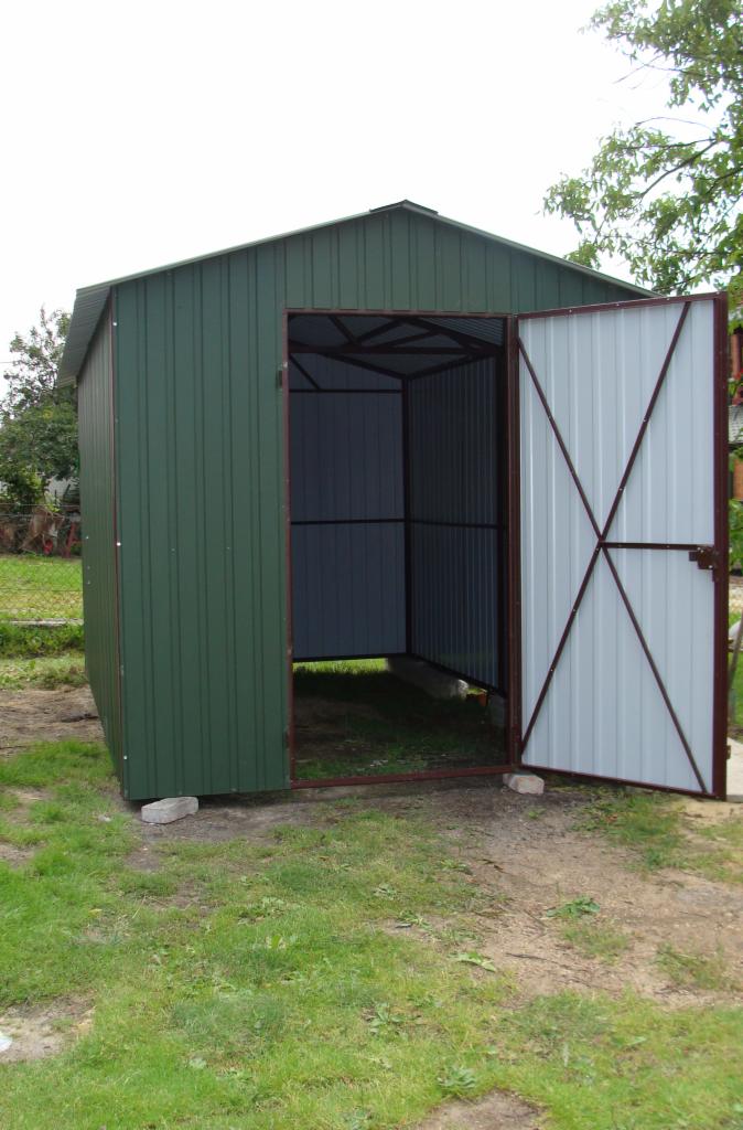 Otwarty, zielony garaż blaszany z białą, częściowo otwartą bramą, osadzony na betonowych bloczkach, na tle zieleni.