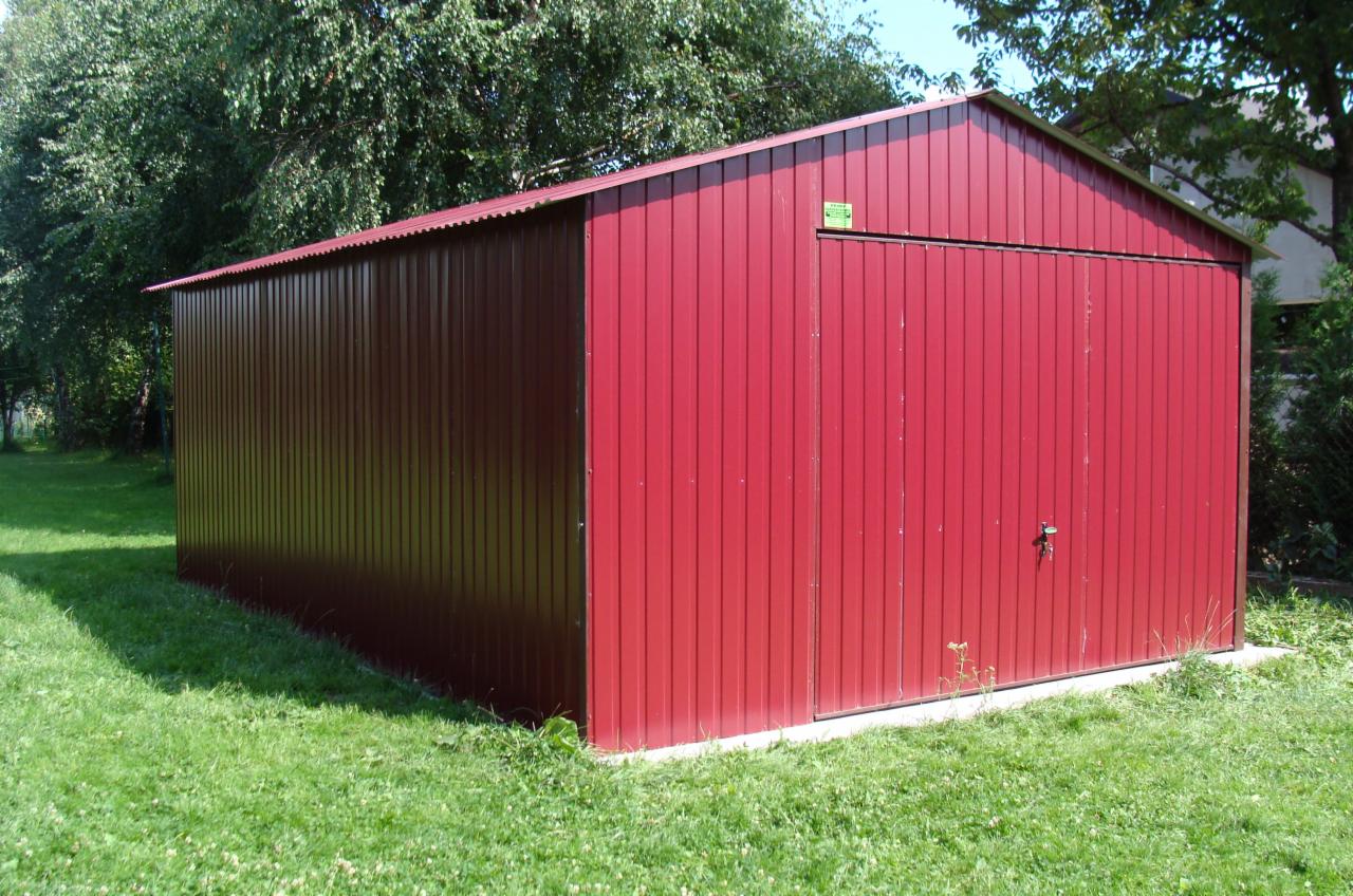 Czerwony garaż blaszany z dwuspadowym dachem, zlokalizowany w Jodłowniku, na trawiastym terenie pod drzewami.