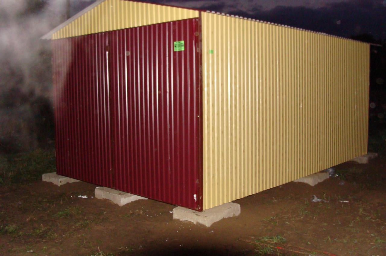 Blaszany garaż w trakcie montażu, z bordową ścianą frontową i beżową boczną, osadzony na betonowych bloczkach na grząskim terenie.