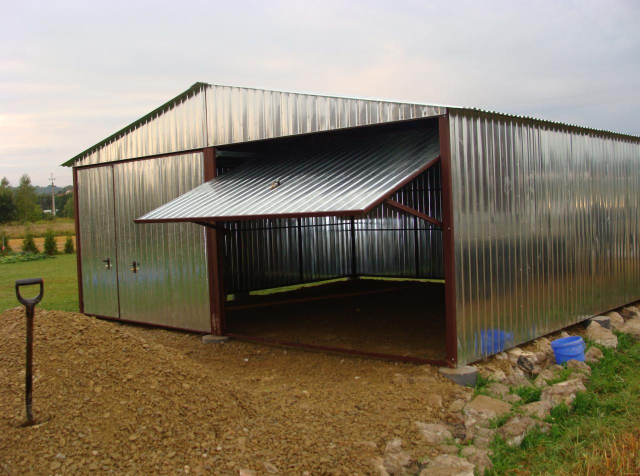 Otwarty, blaszany garaż z dachem dwuspadowym, wykonany z blachy falistej, z brązową ramą, widoczny na tle zieleni i pochmurnego nieba, z łopatą i dwoma niebieskimi wiaderkami w pobliżu.