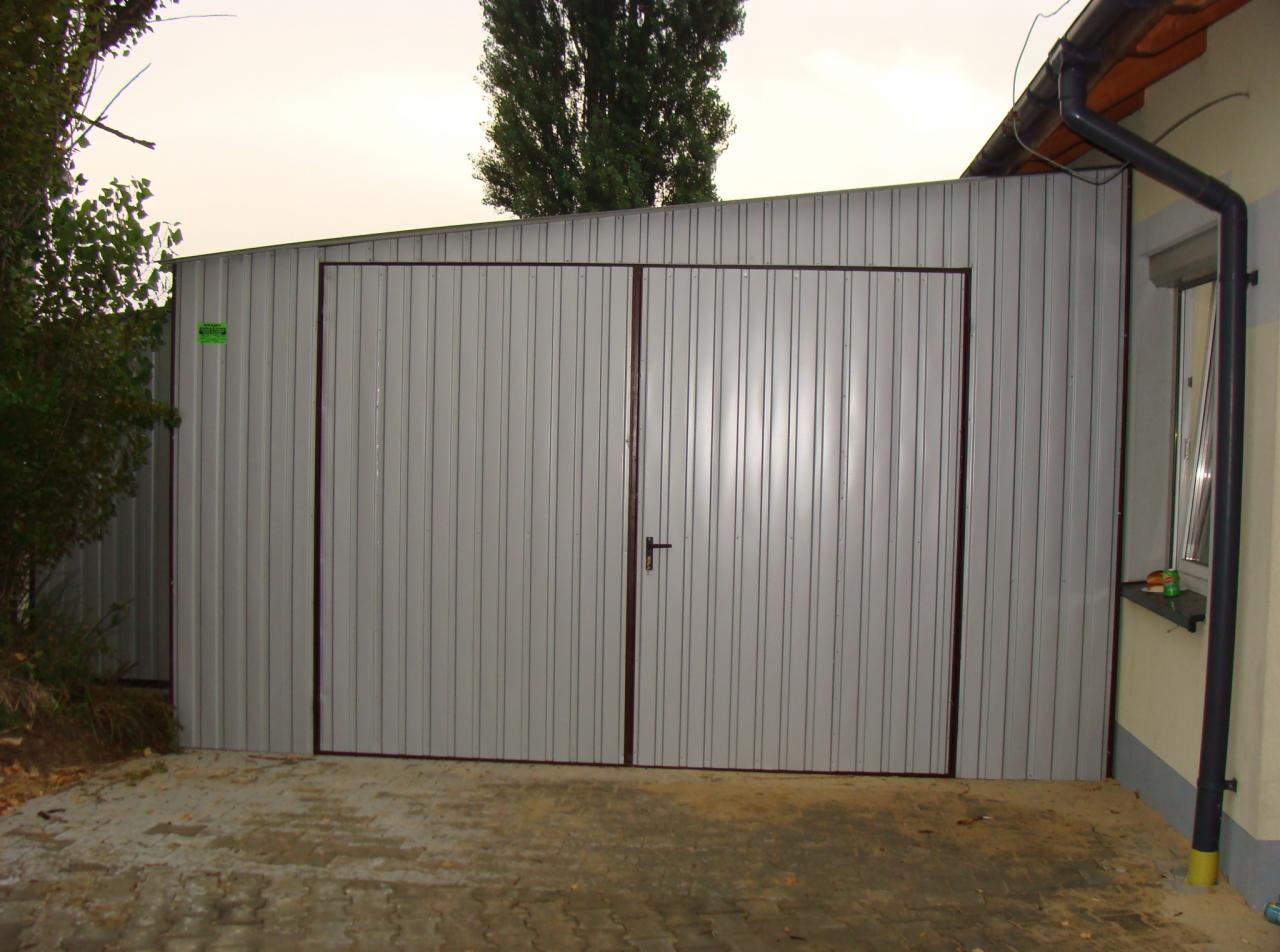 Szary blaszak garażowy z dwuskrzydłowymi drzwiami, obramowanymi brązowymi profilami, stojący na betonowej kostce, z widoczną roślinnością po lewej stronie i fragmentem budynku z rynną po prawej.