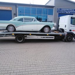 LAWETA Pomoc Drogowa TUCHÓW Transport HOLOWANIE - Klasyczny, dwukolorowy samochód z lat 50. transportowany na platformie pojazdu pomocy drogowej przed budynkiem stacji kontroli pojazdów.