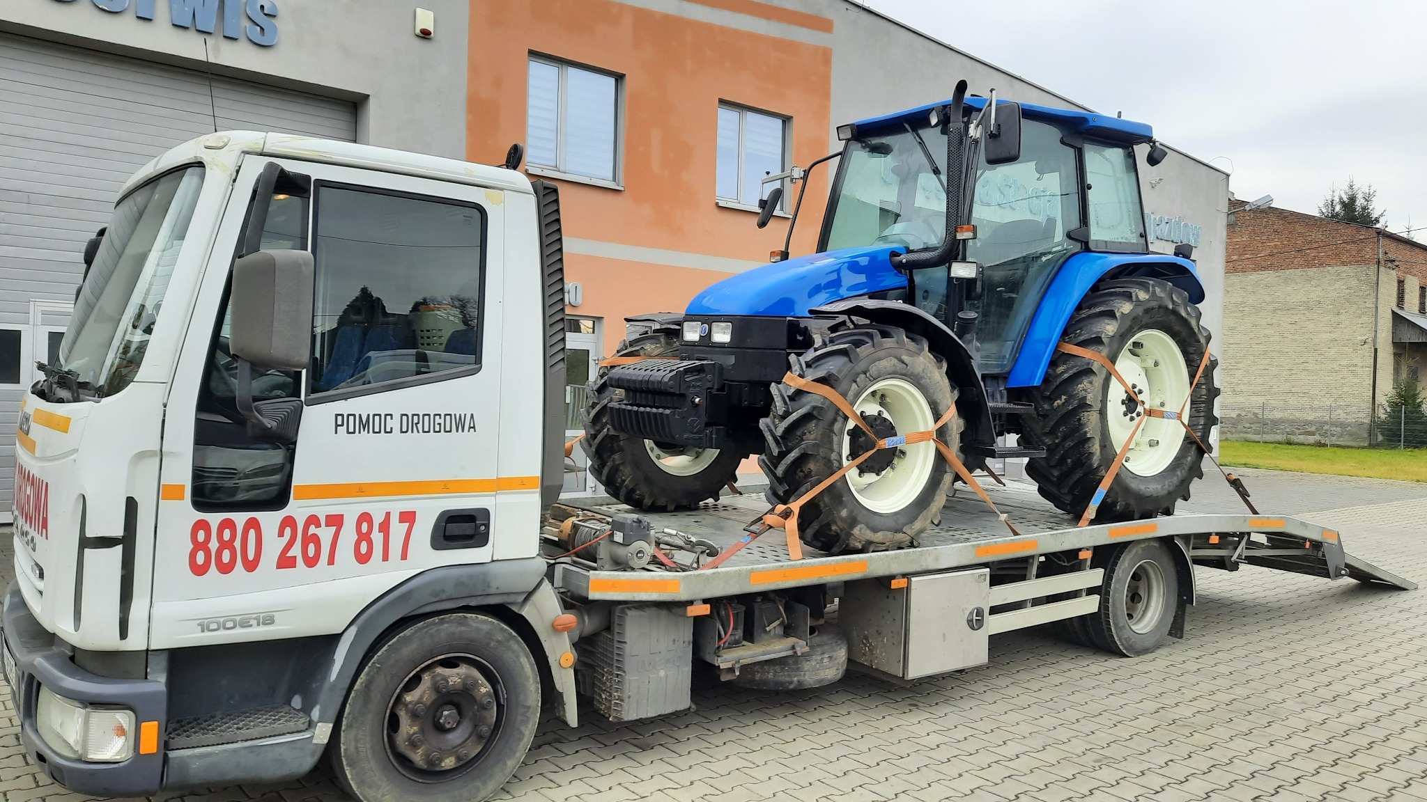 Biała laweta z napisem 'Pomoc Drogowa' przewozi niebieski traktor zabezpieczony pasami transportowymi na tle budynku.