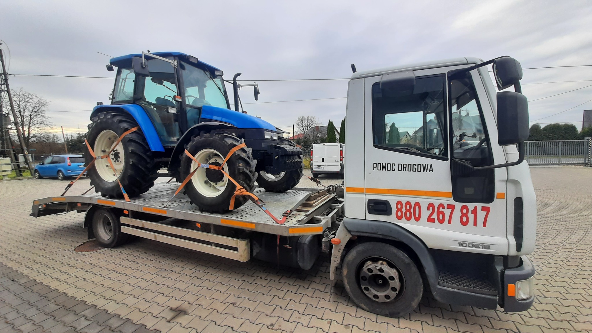 Niebieski traktor zabezpieczony pomarańczowymi pasami na platformie białej ciężarówki z napisem 'POMOC DROGOWA' i numerem telefonu, na tle kostki brukowej i zabudowań.