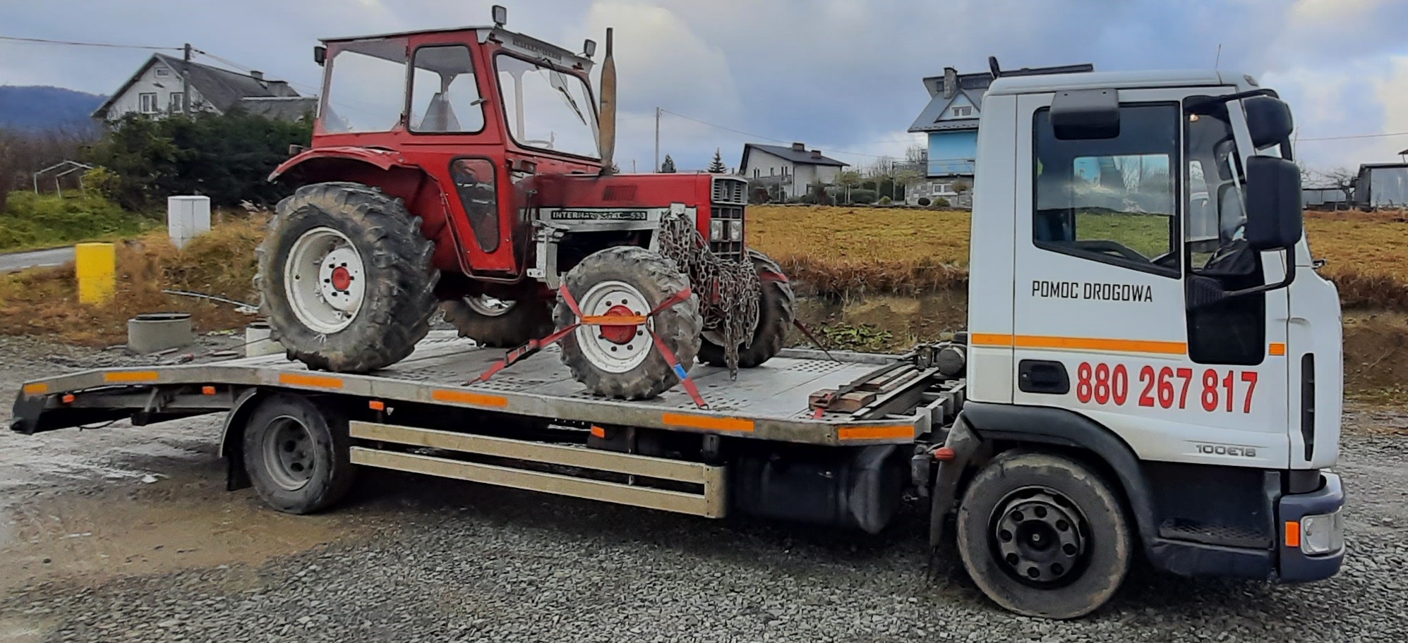 Czerwony traktor International 533 zabezpieczony pasami transportowymi na platformie niskopodwoziowej z napisem 'Pomoc Drogowa' na tle wiejskiego krajobrazu.