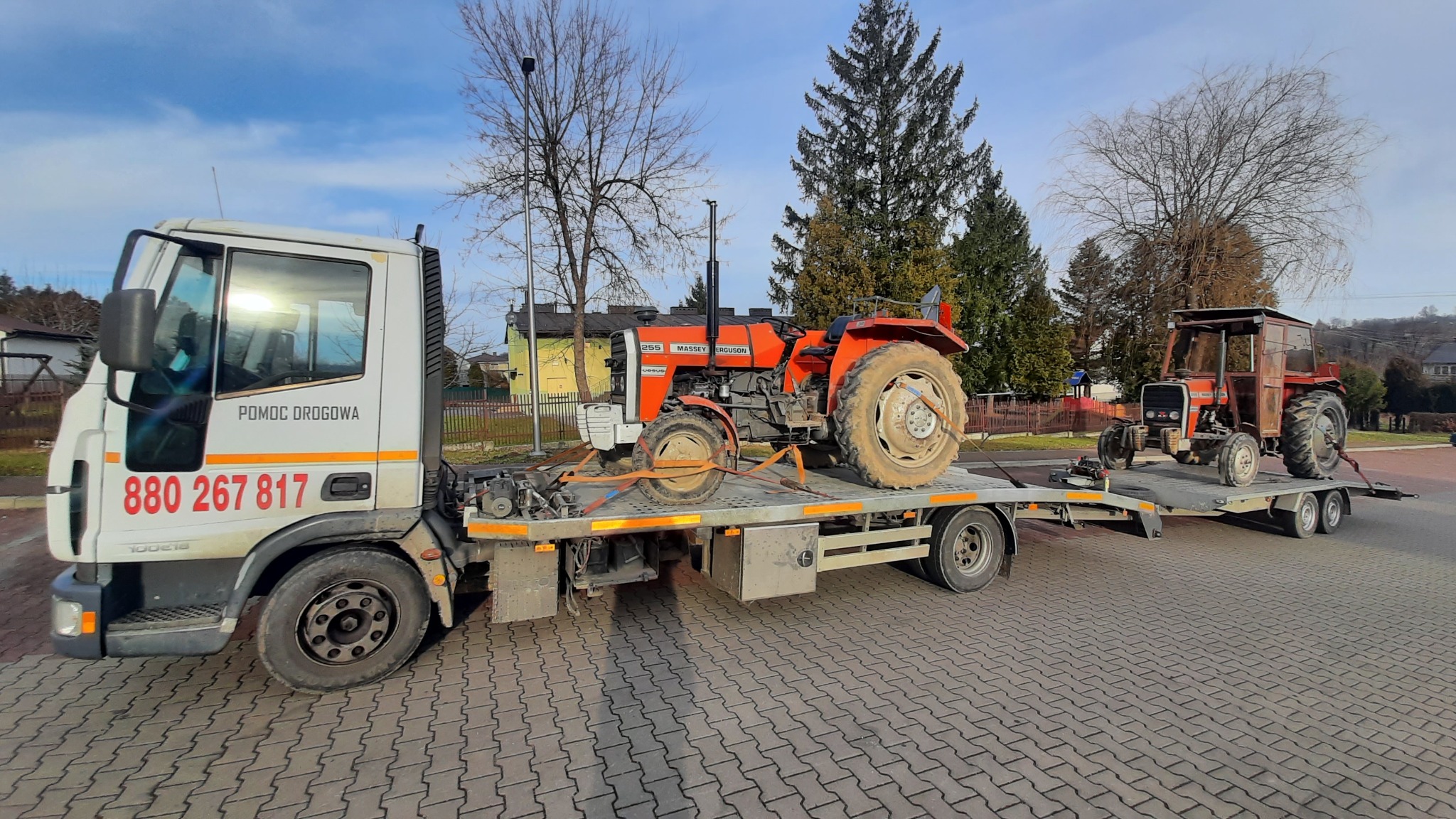Biała ciężarówka pomocy drogowej z dwoma pomarańczowymi traktorami na platformie, zaparkowana na kostce brukowej w słoneczny dzień.