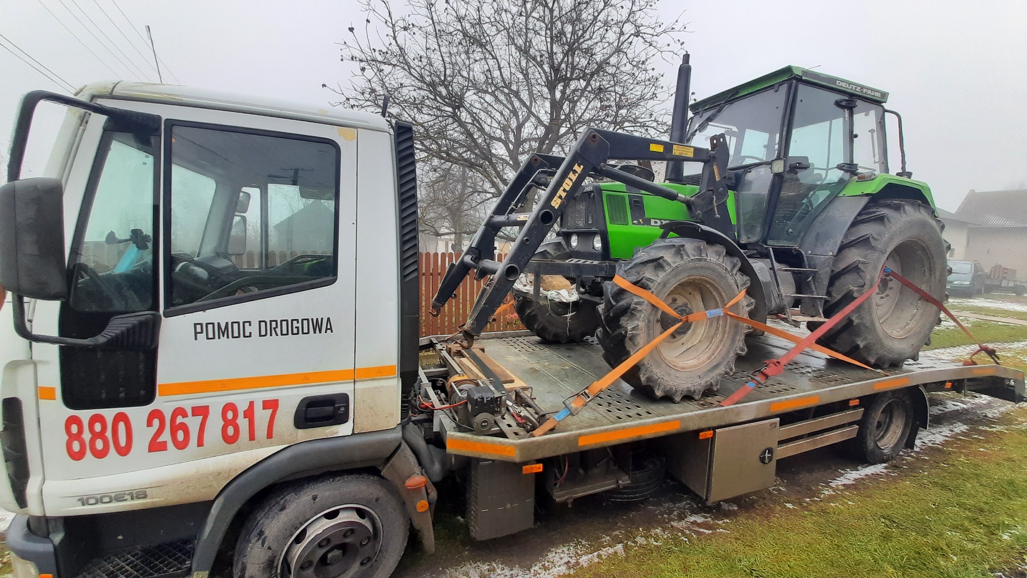 Laweta z napisem 'Pomoc Drogowa' transportuje zielony traktor Deutz-Fahr na platformie zabezpieczonej pomarańczowymi pasami.