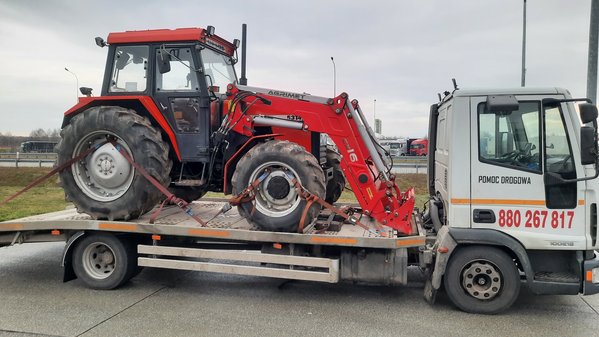 Czerwony traktor z ładowaczem czołowym marki Agrimet 5314, zabezpieczony pasami transportowymi, przewożony na platformie lawety pomocy drogowej z widocznym numerem telefonu.