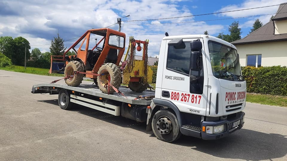 Pomarańczowy traktor rolniczy z kabiną, zabezpieczony pasami transportowymi na białej platformie pojazdu pomocy drogowej z napisem 'Pomoc Drogowa' i numerem telefonu.