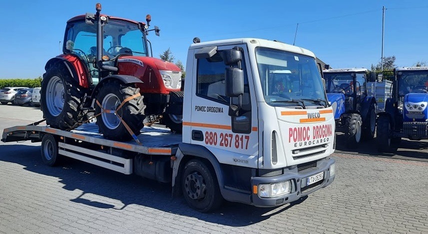 Czerwony traktor zabezpieczony pasami na platformie lawety Iveco Pomoc Drogowa, w tle zaparkowane inne traktory i samochody osobowe.