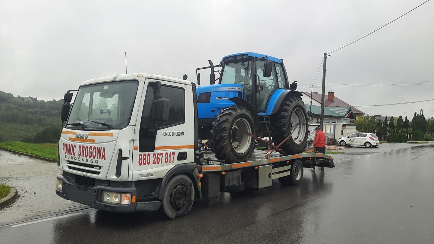 Biała laweta Iveco z napisem 'Pomoc Drogowa Eurocargo' przewozi niebieski traktor Landini Legend 130 na platformie, zabezpieczony pasami transportowymi, na tle wiejskiej drogi i zabudowań.
