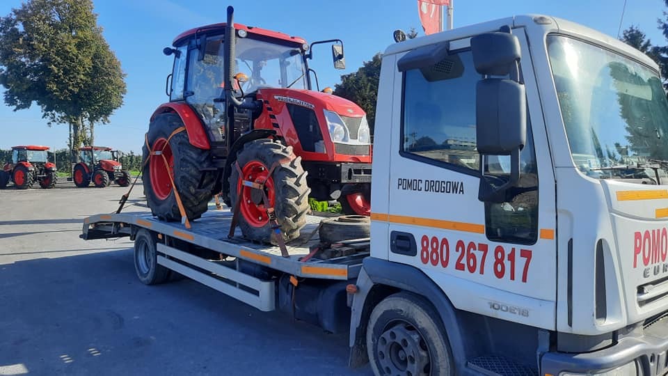 Czerwony traktor Zetor Forterra załadowany na platformę pojazdu pomocy drogowej z widocznym numerem telefonu, w tle inne traktory.
