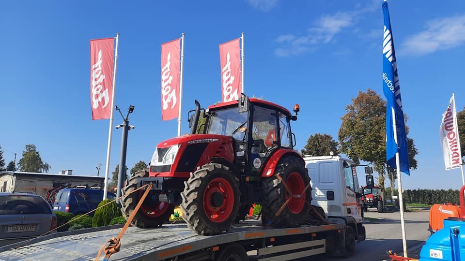 Czerwony traktor Zetor Major CL 80 na lawecie, zabezpieczony pasami transportowymi, w tle budynki i flagi reklamowe.