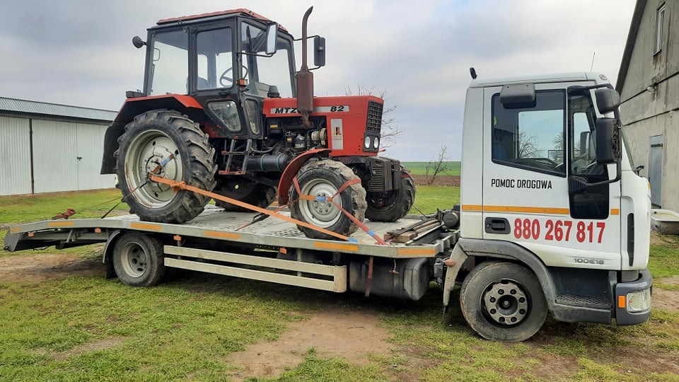 Czerwony traktor MTZ 82 zabezpieczony pasami transportowymi na platformie białej ciężarówki z napisem 'Pomoc Drogowa' na tle wiejskiego krajobrazu.