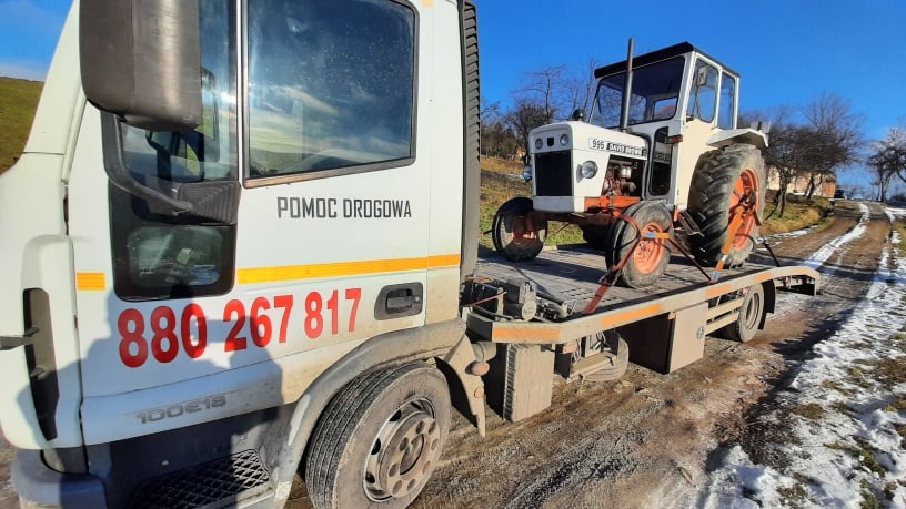 Biała ciężarówka z napisem 'Pomoc Drogowa' i numerem telefonu przewozi na platformie biało-pomarańczowy traktor zabezpieczony pasami transportowymi na wiejskiej drodze z resztkami śniegu.