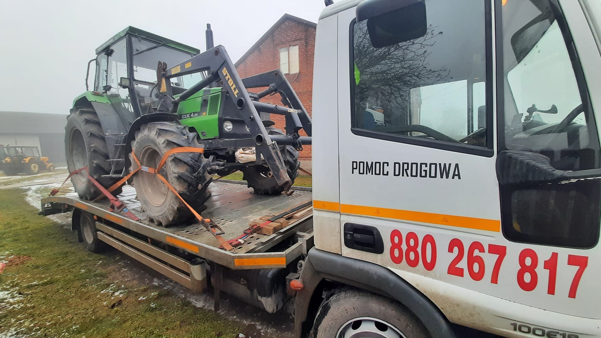 Zielony traktor Deutz-Fahr DX 4.51 z ładowaczem czołowym Stoll, zabezpieczony pomarańczowymi pasami transportowymi na niskopodwoziowej platformie lawety z napisem 'POMOC DROGOWA' i numerem telefonu...