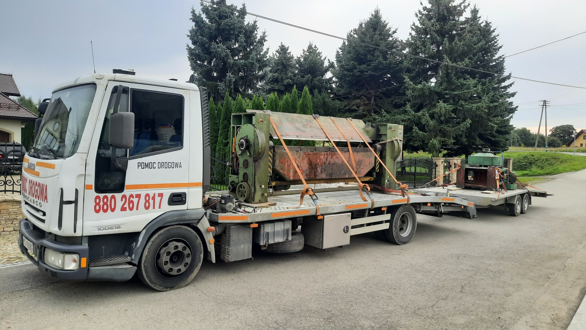 Biała ciężarówka z napisem 'Pomoc Drogowa' przewozi na platformie i przyczepie ciężkie, zielone maszyny przemysłowe, zabezpieczone pomarańczowymi pasami transportowymi.