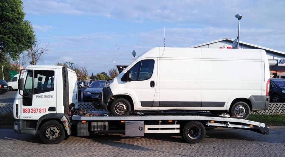 Biała laweta z białym busem na platformie, widoczny numer telefonu 'Pomoc Drogowa' na kabinie, na parkingu przed budynkiem.