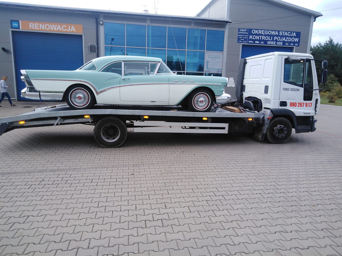 Klasyczny, dwukolorowy samochód z lat 50. transportowany na platformie pojazdu pomocy drogowej przed budynkiem stacji kontroli pojazdów.