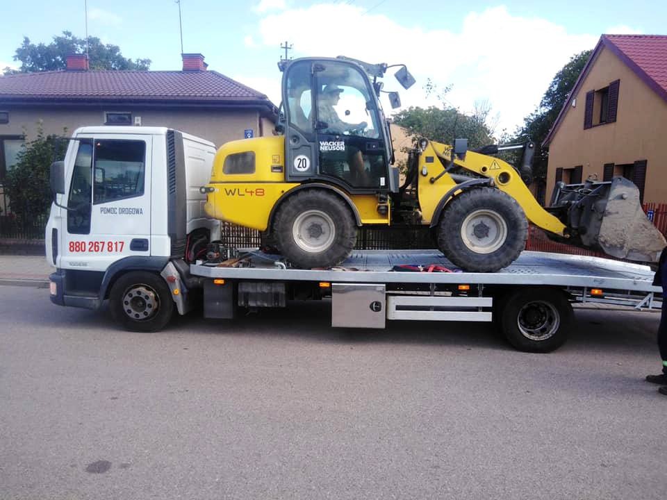 Laweta z Ryglice transportuje żółtą ładowarkę Wacker Neuson WL48 na platformie. Widoczny numer telefonu 'Pomoc Drogowa' na kabinie lawety.