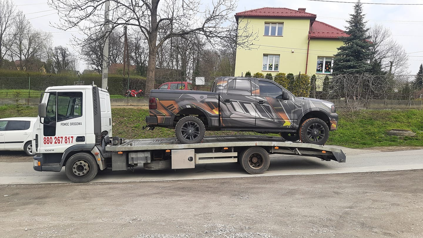 Biała ciężarówka z napisem 'Pomoc Drogowa' przewozi na platformie pickup'a z efektowną, futurystyczną grafiką. W tle żółty dom z czerwonym dachem i drzewa bez liści.