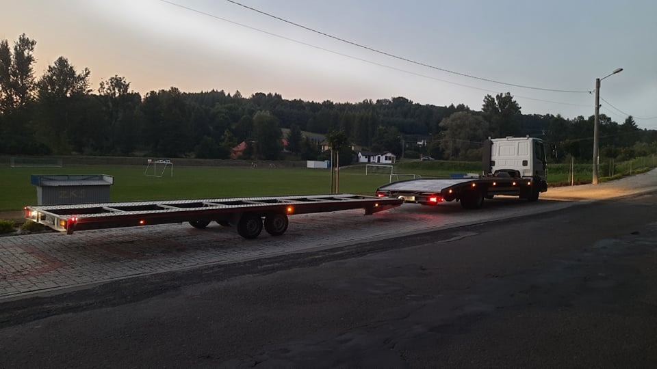 Ciężarówka z platformą do transportu pojazdów, zaparkowana na poboczu drogi, z widocznym boiskiem sportowym w tle, oświetlona światłami pozycyjnymi o zmierzchu.