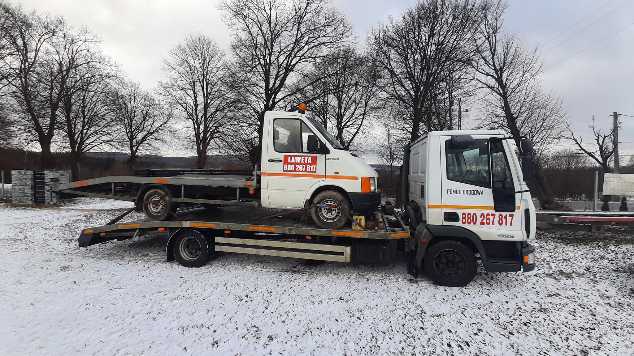 Biała laweta transportuje inny biały samochód dostawczy na tle zimowego krajobrazu z drzewami bez liści.