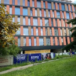 Wielopiętrowy budynek biurowy z elewacją w odcieniach brązu i pomarańczy, częściowo zasłonięty drzewami i ogrodzeniem z reklamami, na szczycie widoczne rusztowania.