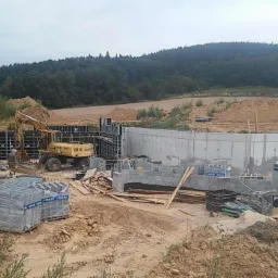 Żółta koparka kołowa ZM 15 przy betonowym fundamencie w trakcie budowy, widoczne szalunki i materiały budowlane, w tle zalesione wzgórze.