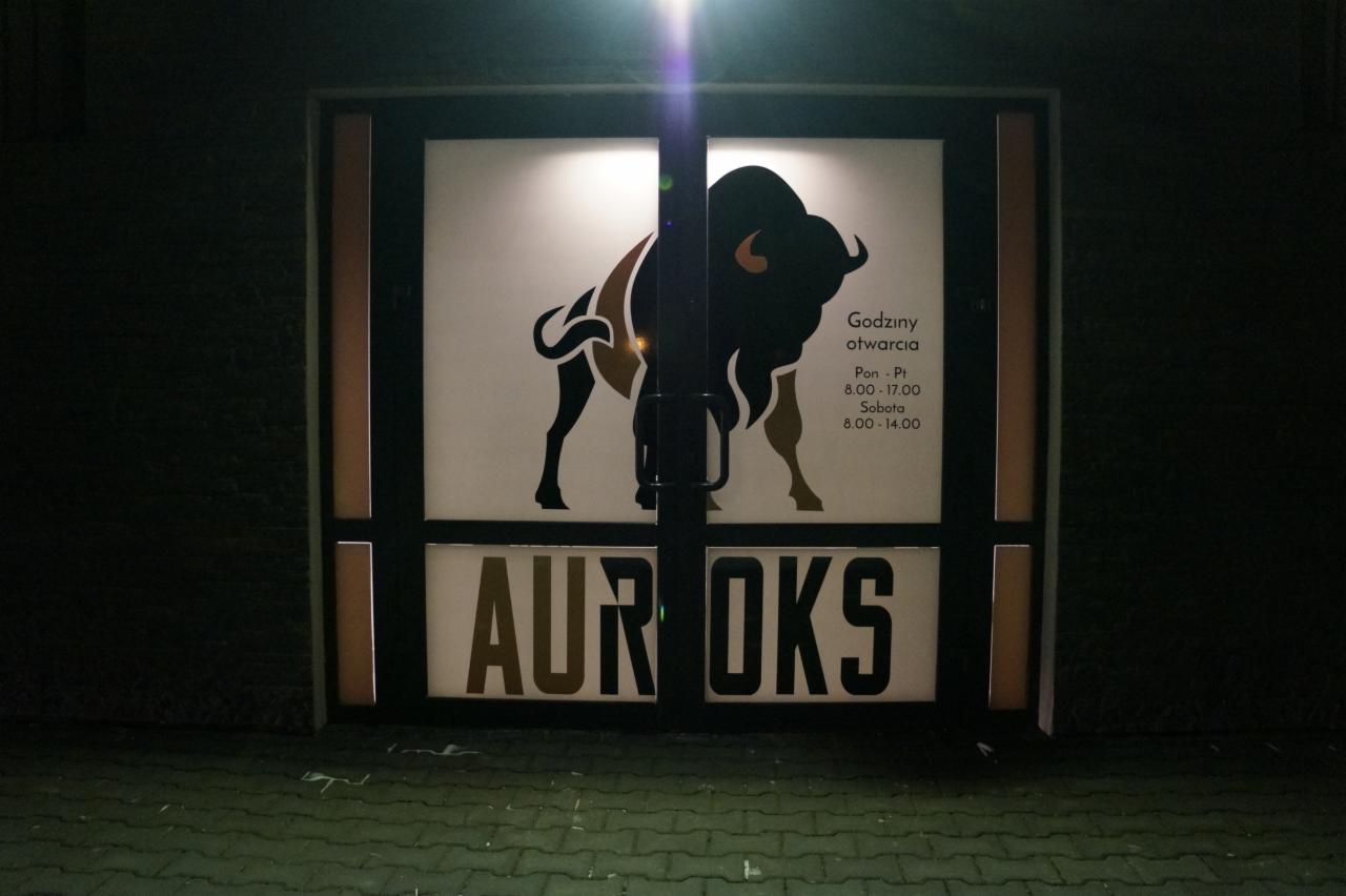 Wejście do sklepu z logo żubra i godzinami otwarcia na szybie, widziane z zewnątrz w słabym oświetleniu.