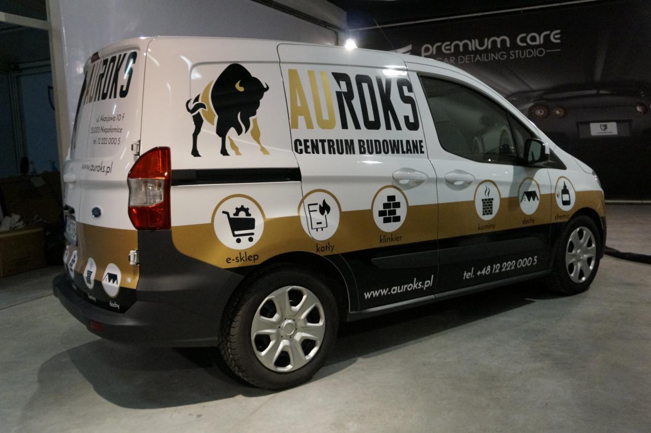 Biały samochód dostawczy z logo firmy budowlanej AUROKS, prezentujący grafikę bizona i ikony e-sklepu, kotłów, klinkieru, kominów, dachów i chemii budowlanej, zaparkowany w pomieszczeniu.