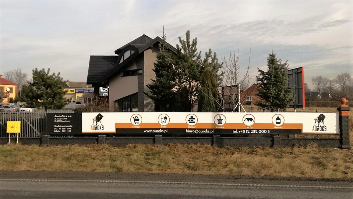Tablica reklamowa firmy Auroks z logotypem bizona, informacjami kontaktowymi i ikonami symbolizującymi: e-sklep, kotły, linkier, kominy, dachy, chemia, na tle budynku z ciemnym dachem i drzewami.
