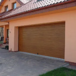 Zewnętrzny widok domu jednorodzinnego z brązową, segmentową bramą garażową o panelach imitujących drewno, z kostki brukowej ułożony podjazd.