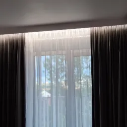 Nowoczesne okno z podwójnymi zasłonami: ciemnoszare, grube zasłony po bokach i białe, prześwitujące firany na środku. Oświetlenie LED ukryte za karniszem podkreśla minimalistyczny design.