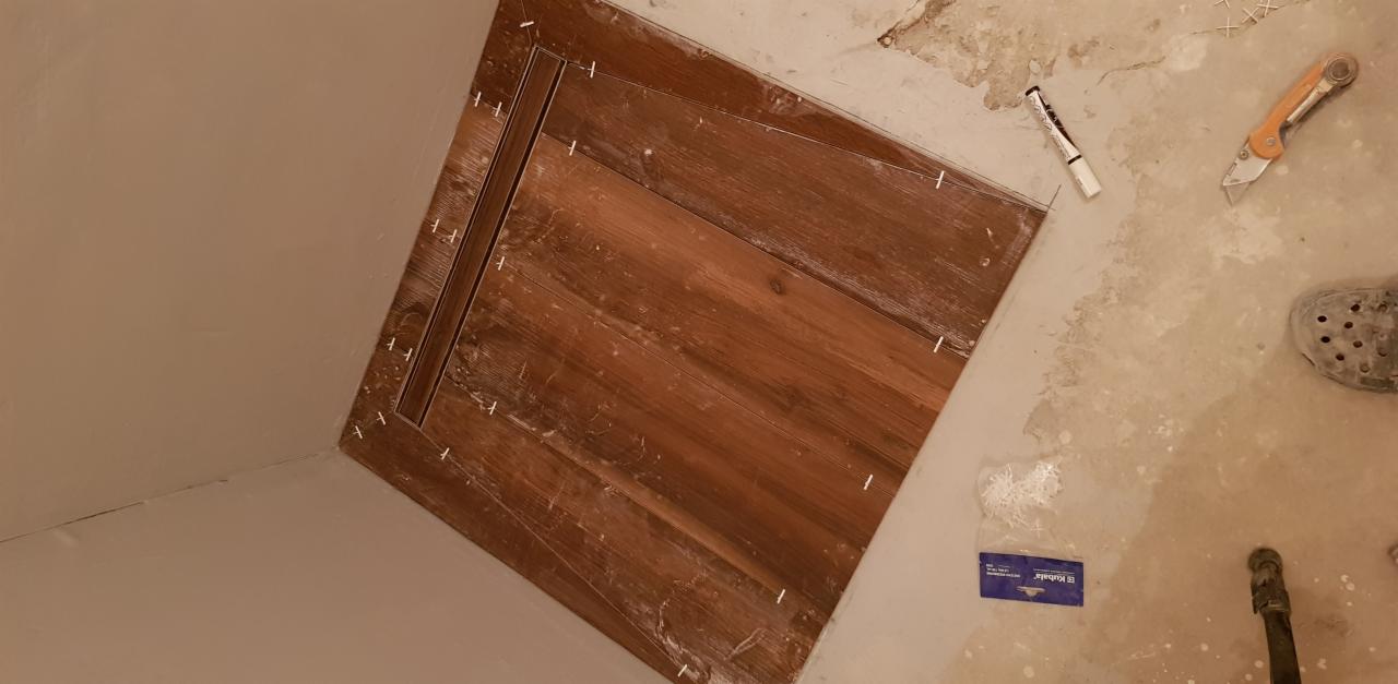 Układanie drewnopodobnych płytek w łazience, widoczny odpływ liniowy, krzyżyki dystansowe, nożyk, marker i fragment stopy w obuwiu roboczym.